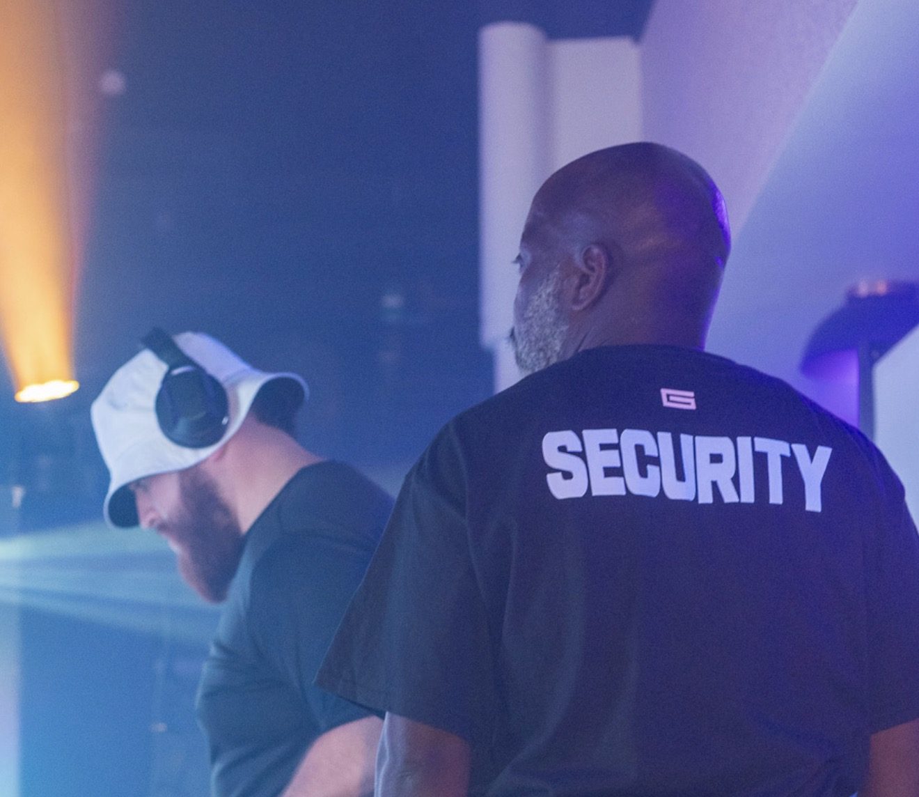 G at the decks in a white bucket hat beside a security guard whose shirt carries the G mark